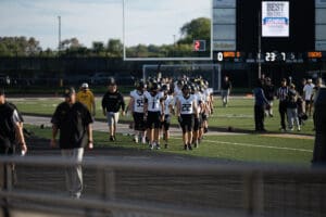 Football team walking onto field under clear sky, nearing scoreboard, in black and white uniforms.