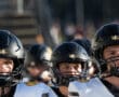 Football players in black helmets and white jerseys enter the field, ready for the game under clear skies.