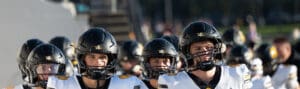 Football players in black helmets and white jerseys enter the field, ready for the game under clear skies.