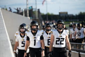 High school football players in black and white uniforms walking onto field, helmets on, ready for the game.