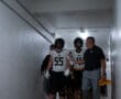 Football players and coach walk through a dim hallway, preparing for a game in uniforms and helmets.