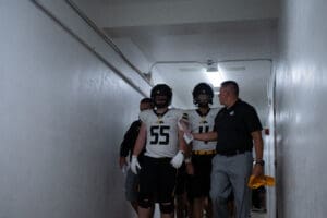 Football players and coach walk through a dim hallway, preparing for a game in uniforms and helmets.