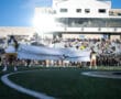 Football players burst through banner held by cheerleaders at stadium entrance, with crowd watching.