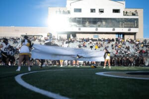 Football players burst through banner held by cheerleaders at stadium entrance, with crowd watching.