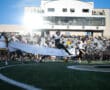 Football player leaps through banner on field as cheerleaders and crowd watch during game day.