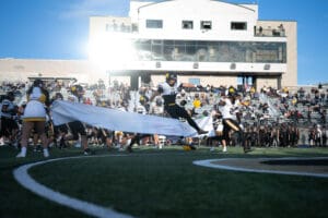 Football player leaps through banner on field as cheerleaders and crowd watch during game day.