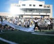 Football players running onto the field with cheerleaders holding a banner, stadium in the background.