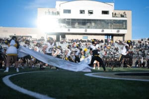 Football players running onto the field with cheerleaders holding a banner, stadium in the background.