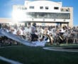 Football team running onto the field through a banner with cheering fans in the stadium background.