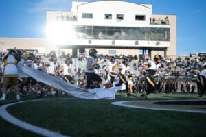 Football team running onto the field through a banner with cheering fans in the stadium background.