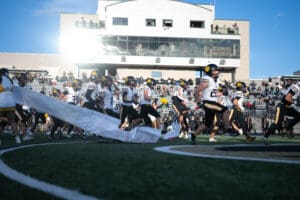 Football team running onto field through banner, energizing crowd in sunny stadium.