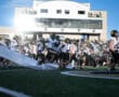 Football team running onto the field during a game, with spectators in the stands under a bright sky.