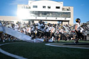 Football team running onto the field during a game, with spectators in the stands under a bright sky.