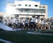 Football team running onto the field in a packed stadium, bright day, crowd in the background, high energy kickoff.