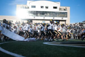 Football team running onto the field in a packed stadium, bright day, crowd in the background, high energy kickoff.