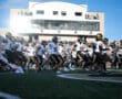 Football team runs onto the field, players wearing black and white uniforms with helmets, crowd in the background.