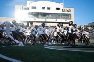 Football team runs onto the field, players wearing black and white uniforms with helmets, crowd in the background.