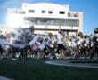 Football team running onto the field, breaking banner, with stadium in the background, captured on a sunny day.