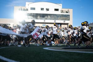 Football team running onto the field, breaking banner, with stadium in the background, captured on a sunny day.