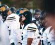 High school football players in white uniforms line up, focused before a game, wearing helmets and sports gear.