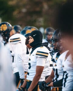 High school football players in white uniforms line up, focused before a game, wearing helmets and sports gear.
