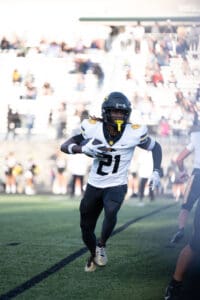 Football player in white and black uniform running with the ball on a field during a game.