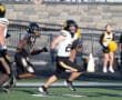 Football player in black and yellow uniform running with ball, pursued by opponent on field with cheerleaders in background.
