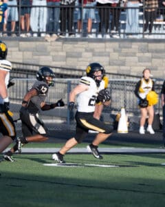 Football player in black and yellow uniform running with ball, pursued by opponent on field with cheerleaders in background.