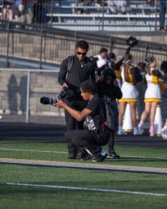 Photographer capturing sports event with telephoto lens on a sunny day at the stadium. Cheerleaders in the background.