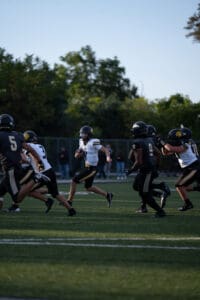Football player running with the ball during a game, teammates and opponents in pursuit on a green field.
