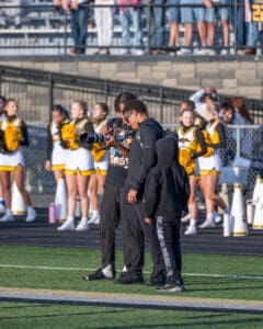Photographers reviewing camera settings on football field, cheerleaders in background.