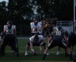 Football players preparing for a snap on a field during a game under evening light.