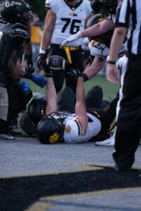 Football player holding ball on the ground during a game, surrounded by teammates and opponents in team jerseys.