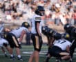 High school football game with teams in action on the field, focused player in white and black gear, crowded stands.