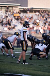 High school football game with teams in action on the field, focused player in white and black gear, crowded stands.