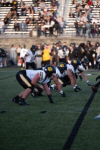 Football players in black and white uniforms line up for the snap on a field during a game.
