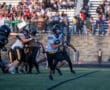 High school football game action with player running the ball past defense on grassy field.