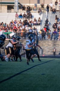 High school football game action with player running the ball past defense on grassy field.