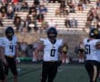 High school football players in helmets and jerseys on the field, with spectators in the background.
