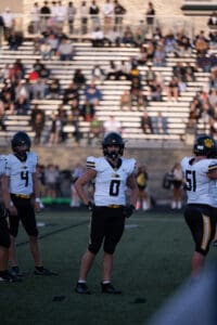 High school football players in helmets and jerseys on the field, with spectators in the background.