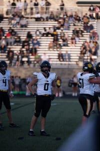 High school football players in uniform on a field during a game, with a crowd in the stadium background.