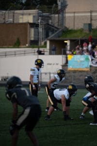High school football game with teams in action, players in white and black uniforms, crowd watching in background.