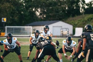 Football game action with players in white jerseys preparing for a snap against opponents in black jerseys.
