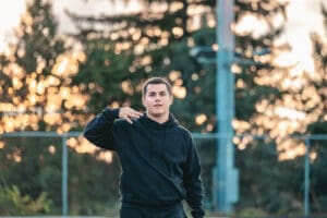 Person in black hoodie outdoors with a blurred nature background at sunset, making a hand gesture.