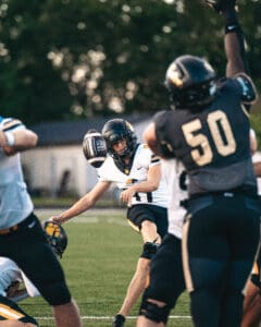 Football player kicks field goal during a game, wearing black and white uniform on a green field.
