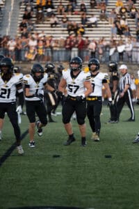 Football players in white jerseys and helmets preparing on the field during a game, with spectators in the stands.