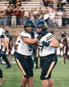 Two football players celebrating on the field with helmets and uniforms, cheering with teammates in the background.