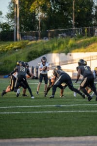 High school football game action with players in motion on the field, green lawn and trees in the background.