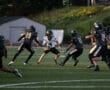 High school football game action, quarterback dodging two defenders on the field at sunset.