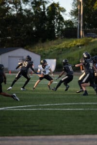 High school football game action, quarterback dodging two defenders on the field at sunset.